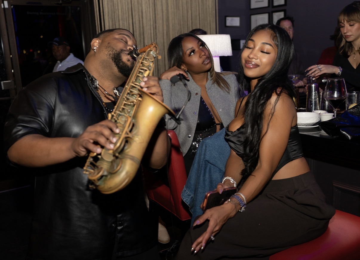Dayve Stewart playing saxophone beside guests at an intimate candlelit lounge table
