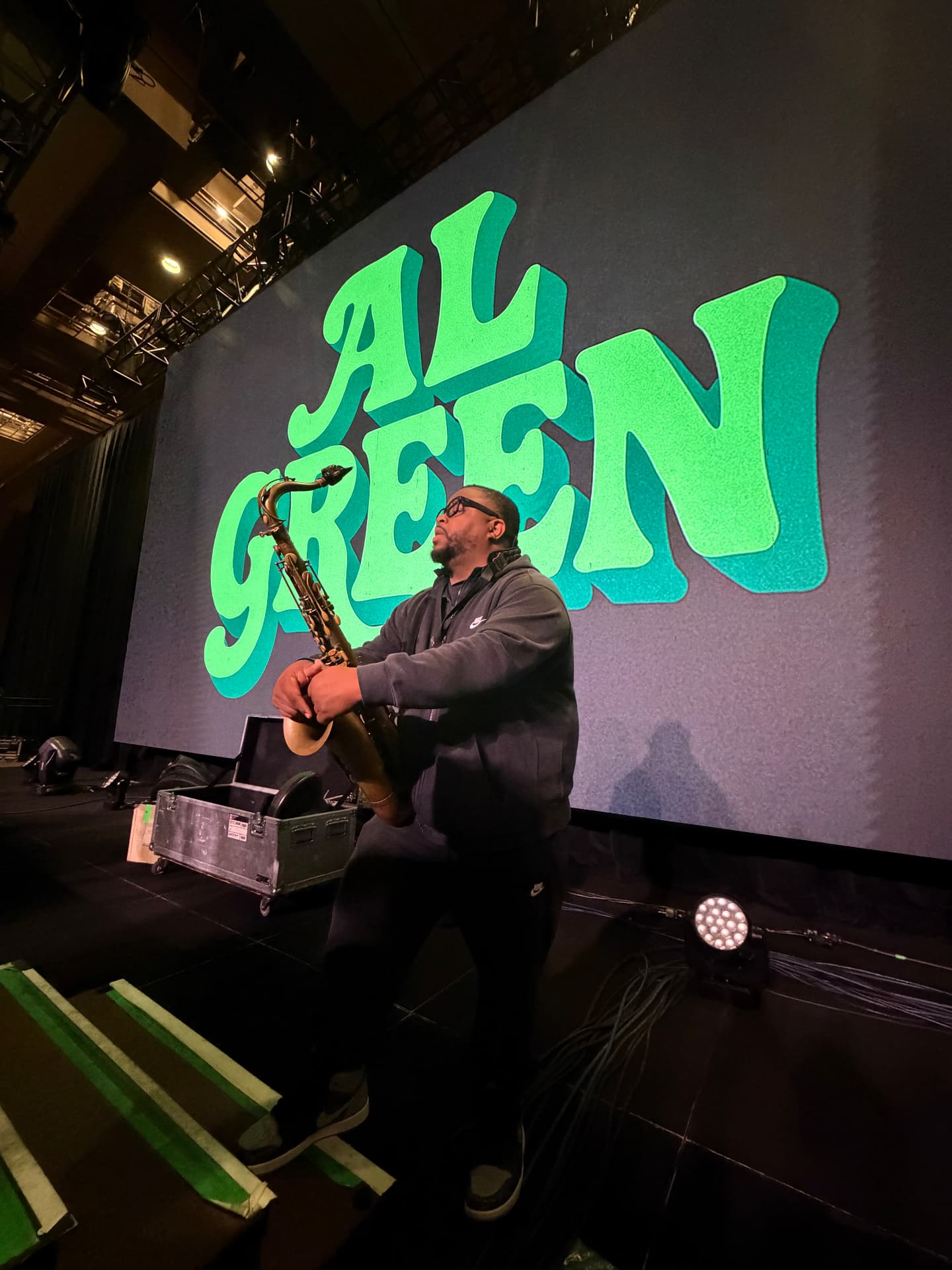 Dayve Stewart holding his tenor saxophone on stage in front of a large Al Green screen