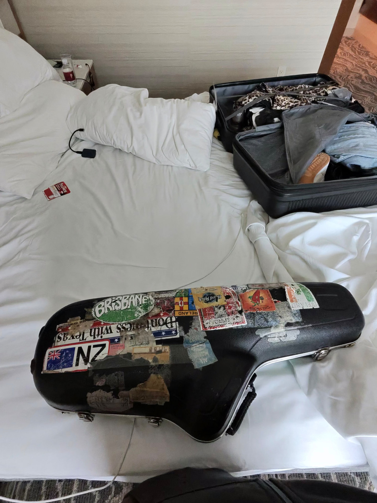 A sticker-covered saxophone case resting on a hotel bed next to an open suitcase